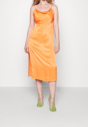 Vrouw in een oranje satijnen midi-jurk met spaghettibandjes en groene sandalen met hoge hakken en bandjes, staand tegen een witte achtergrond.