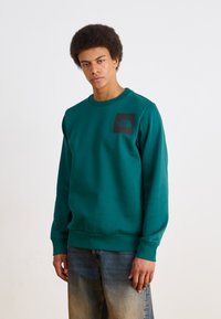 Teal genser laget av en bomullsblandingsstoff, med rund hals, ribbede mansjetter og en svart logo-patch på brystet.