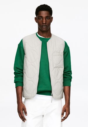 Jeune homme noir aux cheveux courts et bouclés portant une veste matelassée gris clair par-dessus un sweat-shirt vert et un pantalon blanc, sur fond blanc.