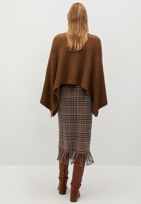 Poncho lavorato a maglia marrone con un design morbido che cade su una gonna plaid lunga fino al ginocchio con dettagli frangiati sull'orlo e stivali marroni alti fino al ginocchio.