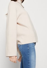 Pull beige à manches longues, col rond, fabriqué en matière douce. Associé à un jean bleu, affichant une coupe décontractée et un design simple.