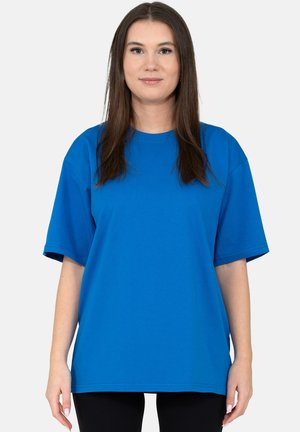 Vrouw met stijlbruin haar die een losse blauwe T-shirt met korte mouwen en een zwarte broek draagt, tegen een effen lichte achtergrond.