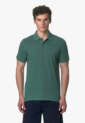 Młody mężczyzna z kręconymi włosami, ubrany w dopasowaną zieloną koszulkę polo i ciemne spodnie, stoi zwrócony przodem na jednolitym tle.