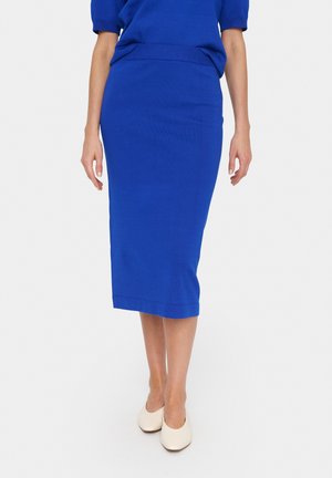 Femme portant une jupe midi bleu roi ajustée, un top assorti et des chaussures blanches fermées à talons bas, debout devant un fond uni.