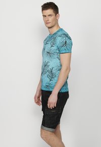 Gaiši zilā T-kreklā ar melniem ziedu un lapu motīviem, apaļš kakla izgriezums, īsas piedurknes, kombinēts ar tumšiem šortiem ar sānu kabatām.