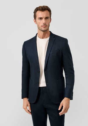 Man in een marineblauwe blazer over een wit T-shirt en bijpassende marineblauwe broek tegen een effen grijze achtergrond.