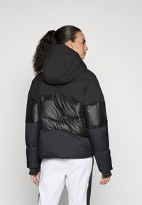 Schwarze, gep padded Jacke mit Kapuze, die eine Mischung aus matten und glänzenden Materialien, gerippten Bündchen und einem Zickzackmuster auf dem Rücken bietet.