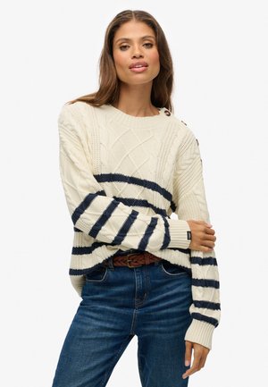 Suéter de punto marfil con franjas azul marino y patrones de diamante texturizados; cuenta con puños acanalados y un cuello redondo, combinado con jeans azules.