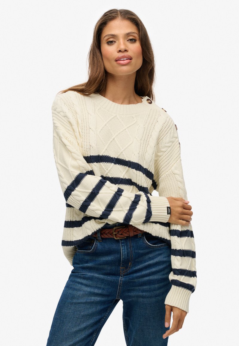 Elfenbeinfarbener Strickpullover mit marineblauen Streifen und strukturierten Rautenmustern; hat gerippte Manschetten und einen Rundhalsausschnitt und wird mit blauen Jeans kombiniert.