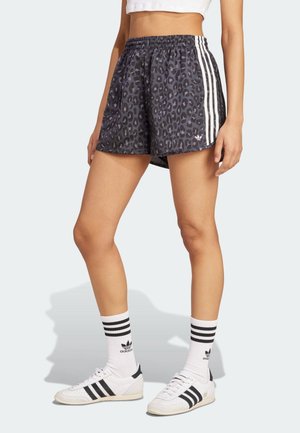 Kobieta ubrana w czarne i szare sportowe szorty w panterkę z białymi paskami, białe skarpetki Adidas oraz białe trampki z czarnymi paskami.