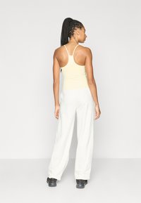 Φωτεινή κίτρινη κοντομάνικη μπλούζα με λεπτές τιράντες, συνδυασμένη με άνετο λευκό παντελόνι. Σκούρα παπούτσια συμπληρώνουν την εμφάνιση. Απλό φόντο.