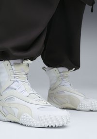 High-top sneakers with a white and light beige color scheme, featuring a textured rubber sole, mesh panels, and intricate lace details.