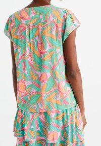 Bluzka w kwiatowy wzór w kolorach zielonego błękitu, różu i pomarańczy. Miękka tkanina z krótkimi rękawami, marszczony detal z tyłu, warstwowy falbaniasty dół.