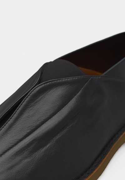 Chaussure à enfiler en cuir noir à texture lisse avec une semelle marron, présentée en gros plan sur un fond clair.