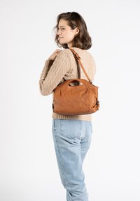 Tasche aus braunem Leder mit weicher Textur, einzigartigem, gebogenem Griff und verstellbarem Schultergurt. Über der Schulter getragen vor einem hellen Hintergrund.