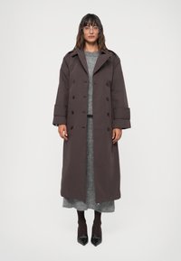 Hnedý dlhý kabát s dvojradovým zapínaním, veľkým golierom a širokými rukávmi. Nosený cez šedý vrstvený outfit, doplnený čiernymi špicatými topánkami.