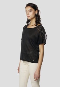 Fekete rövid ujjú felső díszítő mintákkal a vállakon, laza szabású, puha tapintású, világos színű nadrággal párosítva.