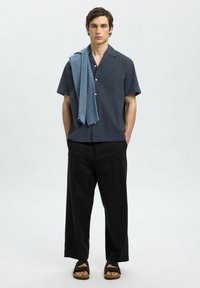 Kortermet, marineblå teksturert skjorte, svarte vide bukser, blå håndkle drapert over skulderen, brune sandaler med flatt såle.