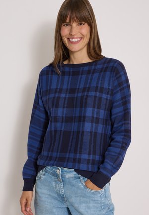 Frau lächelnd, trägt einen blau-schwarz karierten Langarmpullover und hellblaue Jeans mit einer Hand in der Tasche vor einfachem Hintergrund.