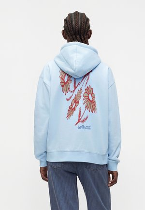 Person med flettet hår iført lys blå hættetrøje med stort rødt og beige blomsterdesign og teksten "Santa Cruz" på ryggen.