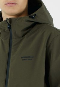 Giacca verde oliva con tessuto dalla texture liscia, cerniera frontale, cappuccio regolabile e logo con la scritta "Refrigiwear Since 1954".