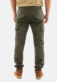 Pantalons cargo vert olive avec des poignets élastiques, plusieurs poches et une texture lisse. Portés avec des bottes marron au revêtement texturé.