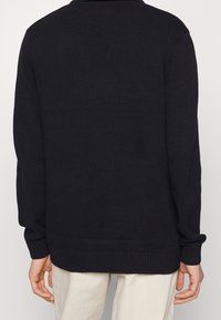 Maglione nero lavorato a maglia, texture a costine, scollo rotondo, maniche lunghe, fessure laterali, vestibilità rilassata, lunghezza media, senza loghi o motivi visibili.