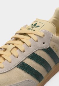 Κοντινό πλάνο σε μπεζ αθλητικό παπούτσι Adidas Samba με σκούρες πράσινες ρίγες και κρεμ κορδόνια, που αναδεικνύει την υφή από σουέντ και δέρμα.