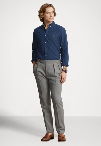 Navy button-down shirt with a small red logo, paired with light gray pleated trousers and brown leather loafers. Smooth fabric textures.