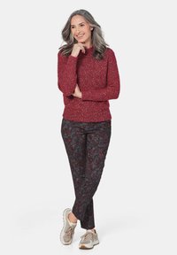 Rot gepunkteter Strickpullover mit hohem Kragen, kombiniert mit dunkel gemusterten Blumenhosen und hellen Sneakers. Model steht in einer entspannten Pose.