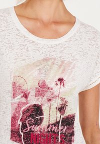 Vit T-shirt med kort ärm och en texturerad mönster. Har en flerfärgad grafisk tryck av palmträd och texten "Sommarkvällar" i röd.