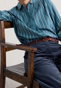 Person wearing teal and white striped button-up shirt, brown woven belt, and dark blue corduroy pants sitting on wooden chair.