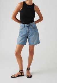 Shorts vaqueros en lavado azul claro con cintura alta, que cuentan con cinco bolsillos y corte de pierna recta. Combinados con un top sin mangas de canalé negro y sandalias negras.