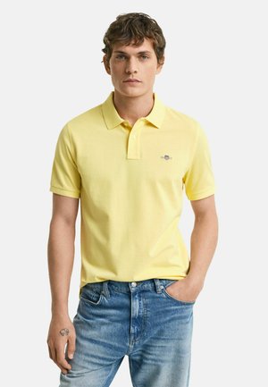 Polo giallo in cotone, con colletto a tre bottoni, maniche corte e un piccolo logo ricamato sul petto. Indossato con jeans blu.