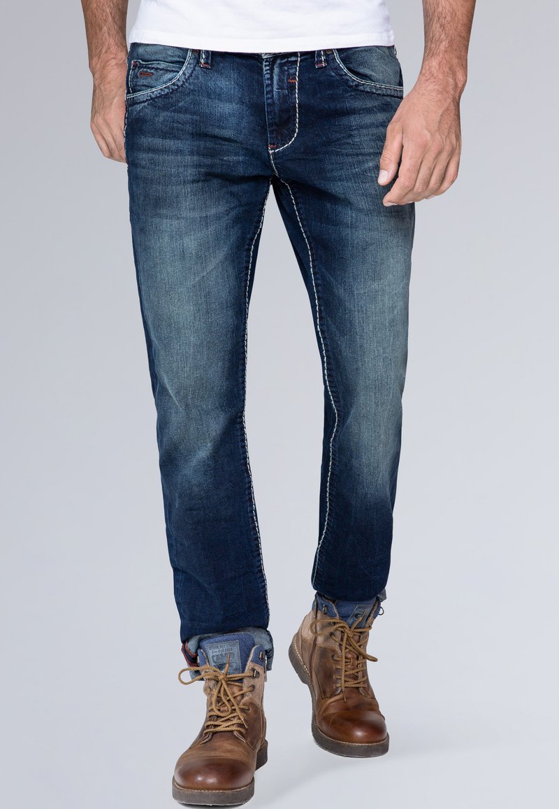 Jeans in denim blu scuro con leggere scoloriture, design a cinque tasche, cuciture a contrasto e vestibilità rilassata. Abbinati a stivali marroni con lacci.
