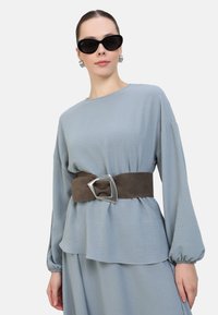 Hellblaues Oberteil mit langen Ärmeln, strukturiertem Stoff, geschichtetem Design, gesichert mit einem braunen Wildledergürtel mit einer silbernen, eckigen Schnalle.