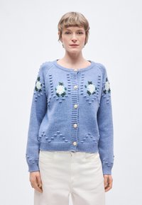 Γαλάζια πλεκτή ζακέτα με κεντήματα λουλουδιών, στρογγυλή λαιμόκοψη και διακοσμητικά κουμπιά. Υφή με στρογγυλά στοιχεία και ριμπ μανσέτες προσθέτουν λεπτομέρεια.