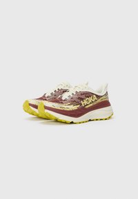 Trailrunning-Schuhe in Bordeauxrot und Creme mit gelben Akzenten. Das Material verfügt über ein strukturiertes Mesh-Obermaterial und eine dicke, gepolsterte Sohle für Stabilität.