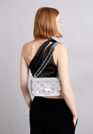Femme aux cheveux rouges courts portant un top noir à une épaule et un petit sac à main argenté à chaîne sur le dos.