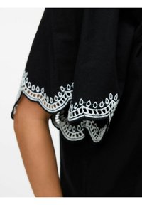 Indumento nero con una delicata bordatura in pizzo ricamato bianco a contrasto sulle maniche, che presenta intricati motivi e una consistenza morbida.