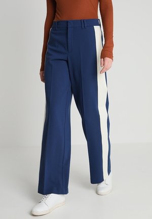 Femme portant un pantalon large bleu marine avec des bandes blanches sur les côtés, associé à des baskets blanches et un haut à manches longues côtelé de couleur rouille.