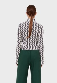 Strickoberteil mit einem wellenförmigen schwarz-weißen Muster, das einen hohen Kragen und lange Ärmel aufweist. Kombiniert mit hochgeschnittenen grünen Hosen.