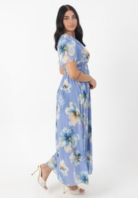 Florales Maxikleid aus leichtem chiffon in Hellblau, mit großen weißen und blauen Blumen, kurzen Ärmeln und tailliertem Schnitt. Kombiniert mit weißen, hohen Sandalen.