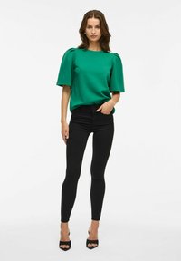 Top verde de manga corta con hombros abullonados, combinado con jeans skinny negros y tacones abiertos negros. Textura suave y diseño ajustado.