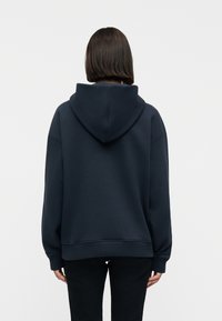 Άτομο με κοντά σκουρόχρωμα μαλλιά που φοράει μια φαρδιά σκουρόχρωμη μπλούζα με κουκούλα και μαύρο παντελόνι, γυρισμένο προς τα πίσω σε απλό φωτεινό φόντο.