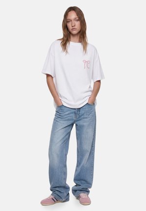 Jeune femme debout, les mains dans les poches, portant un jean bleu clair ample, un T-shirt blanc oversize avec un logo rose et des baskets roses.