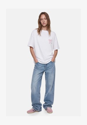 Jeune femme debout, les mains dans les poches, portant un jean bleu clair ample, un T-shirt blanc oversize avec un logo rose et des baskets roses.