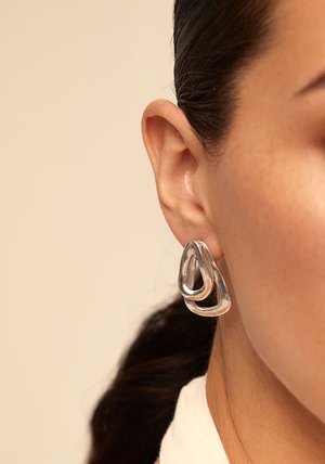 Boucles d'oreilles métalliques argentées présentant des formes de gouttes entrelacées avec une finition polie, suspendues au lobe de l'oreille contre des cheveux mi-longs.