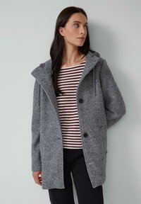 Manteau gris en mélange de laine avec une capuche, présentant une finition texturée, deux boutons noirs et une coupe décontractée, porté sur un haut rayé rouge et blanc.