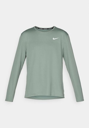 Ilgų rankovių sportinis marškinėliai, pagaminti iš matinės žalios medžiagos, su apvaliu iškirpimu ir baltu Nike logotipu kairėje krūtinės pusėje. Lygi tekstūra.
